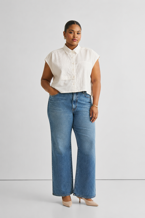Full-length front view of a plus-size model wearing the Reistor Oversized Embroidered Panel Crop Top in White. The organic cotton button-down shirt features distinct texturized embroidered panels and cap sleeves, paired with classic blue wide-leg jeans.