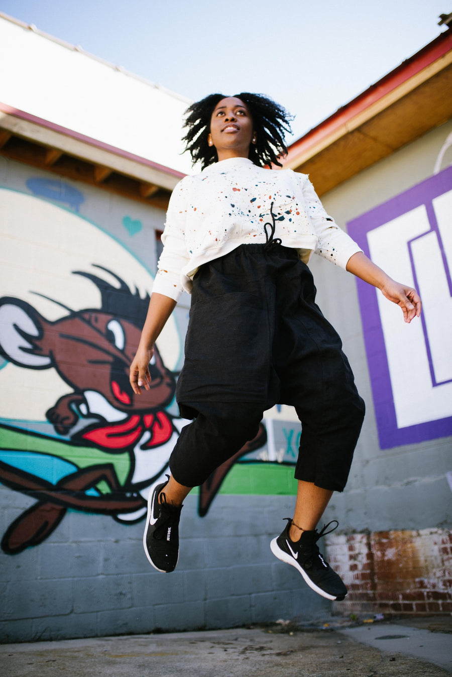 A model wearing the white Megan-Ilene Hand Painted & Quilted Organic Hemp ScrapShirt with colorful splatters and black Puzzle Pants, jumping against a colorful graffiti wall background