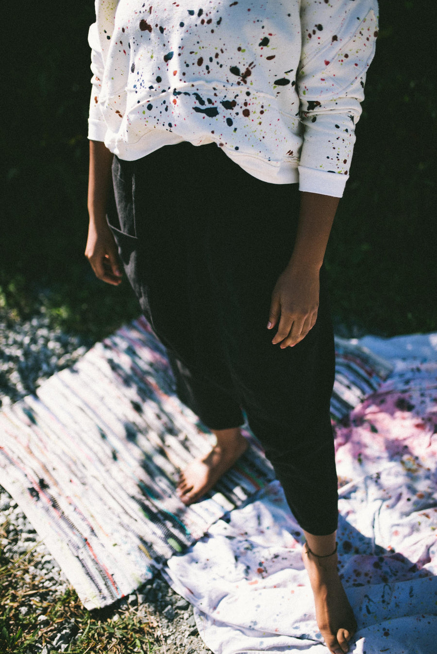 Lower angle view highlighting the black Puzzle Pants paired with the white hand-painted ScrapShirt and the model's bare feet standing on an outdoor mat