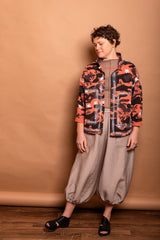 Full-length studio shot of a model wearing the Trompe-l'œil Studio Jacket in vibrant Sherbet orange and black camouflage paired with beige wide-leg trousers.
