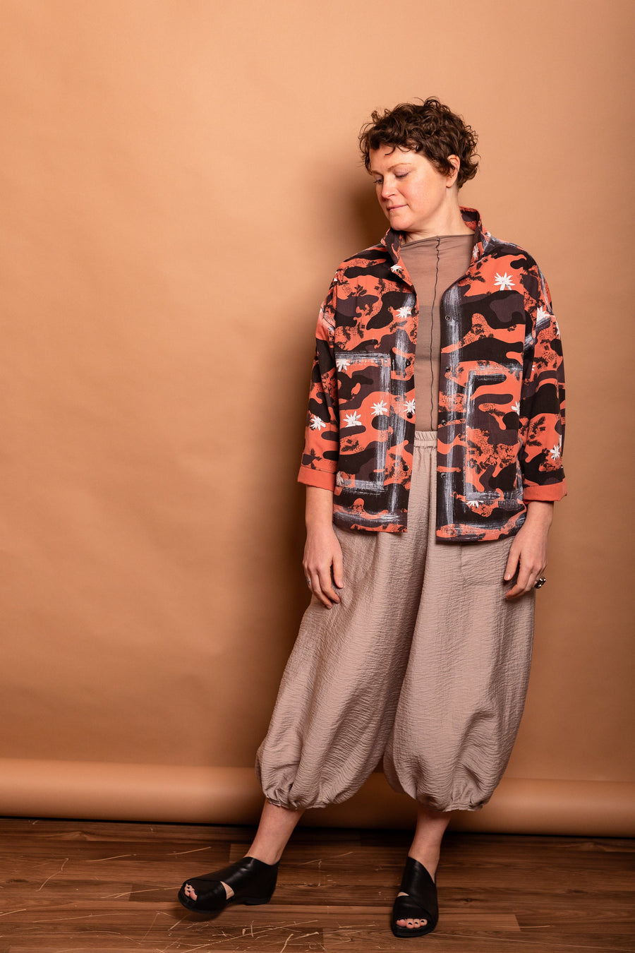Full-length studio shot of a model wearing the Trompe-l'œil Studio Jacket in vibrant Sherbet orange and black camouflage paired with beige wide-leg trousers.