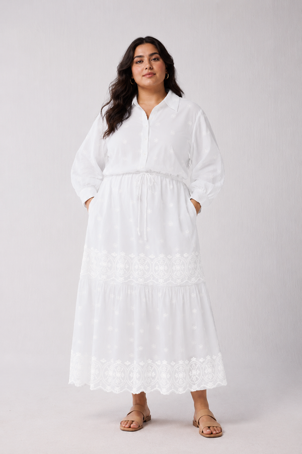 Full-length front view of a plus-size model wearing Reistor's Embroidered Flowy Maxi Tiered Dress in White. The organic cotton dress features a straight collar, front button placket, drawstring waist, and bishop sleeves, styled with tan slide sandals.