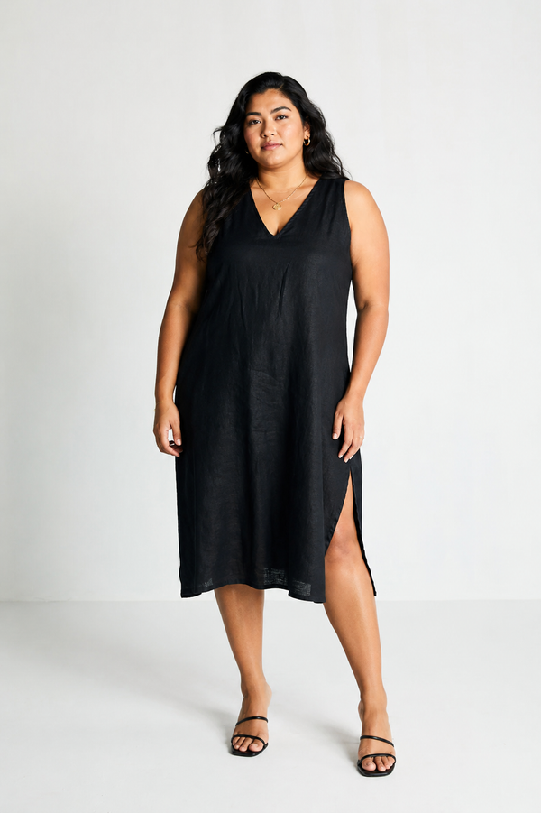 Full-length front view of a plus-size model wearing Reistor's The Hemp Noir Dress. The sleeveless black midi dress is crafted from sustainable 100% hemp fabric and features a sharp V-neckline and high side slits.
