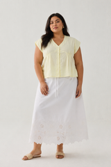 Full-length front view of a plus-size model wearing Reistor's Deep V Neck Top in Butter Lemon. The pale yellow linen blouse features short cap sleeves and a button-down front, styled with a long white embroidered eyelet skirt against a plain light background.