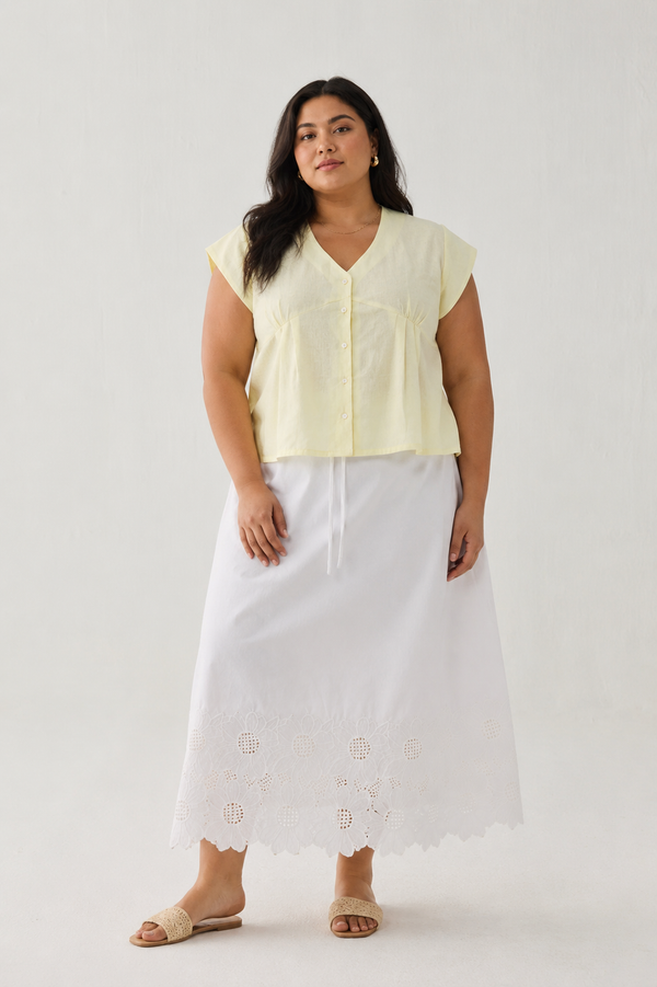 Full-length front view of a plus-size model wearing Reistor's Deep V Neck Top in Butter Lemon. The pale yellow linen blouse features short cap sleeves and a button-down front, styled with a long white embroidered eyelet skirt against a plain light background.