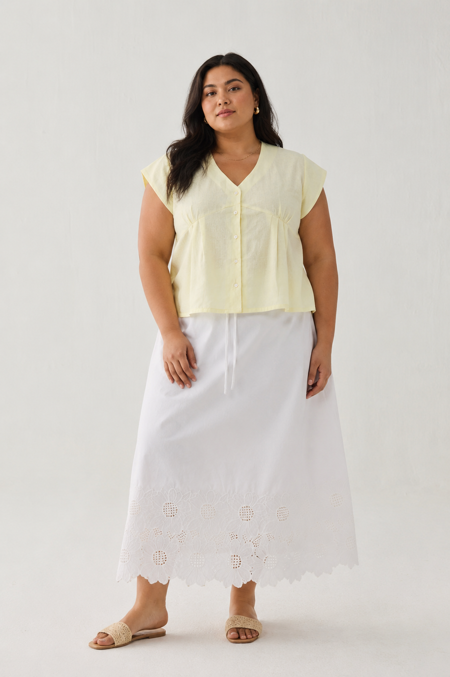 Full-length front view of a plus-size model wearing Reistor's Deep V Neck Top in Butter Lemon. The pale yellow linen blouse features short cap sleeves and a button-down front, styled with a long white embroidered eyelet skirt against a plain light background.