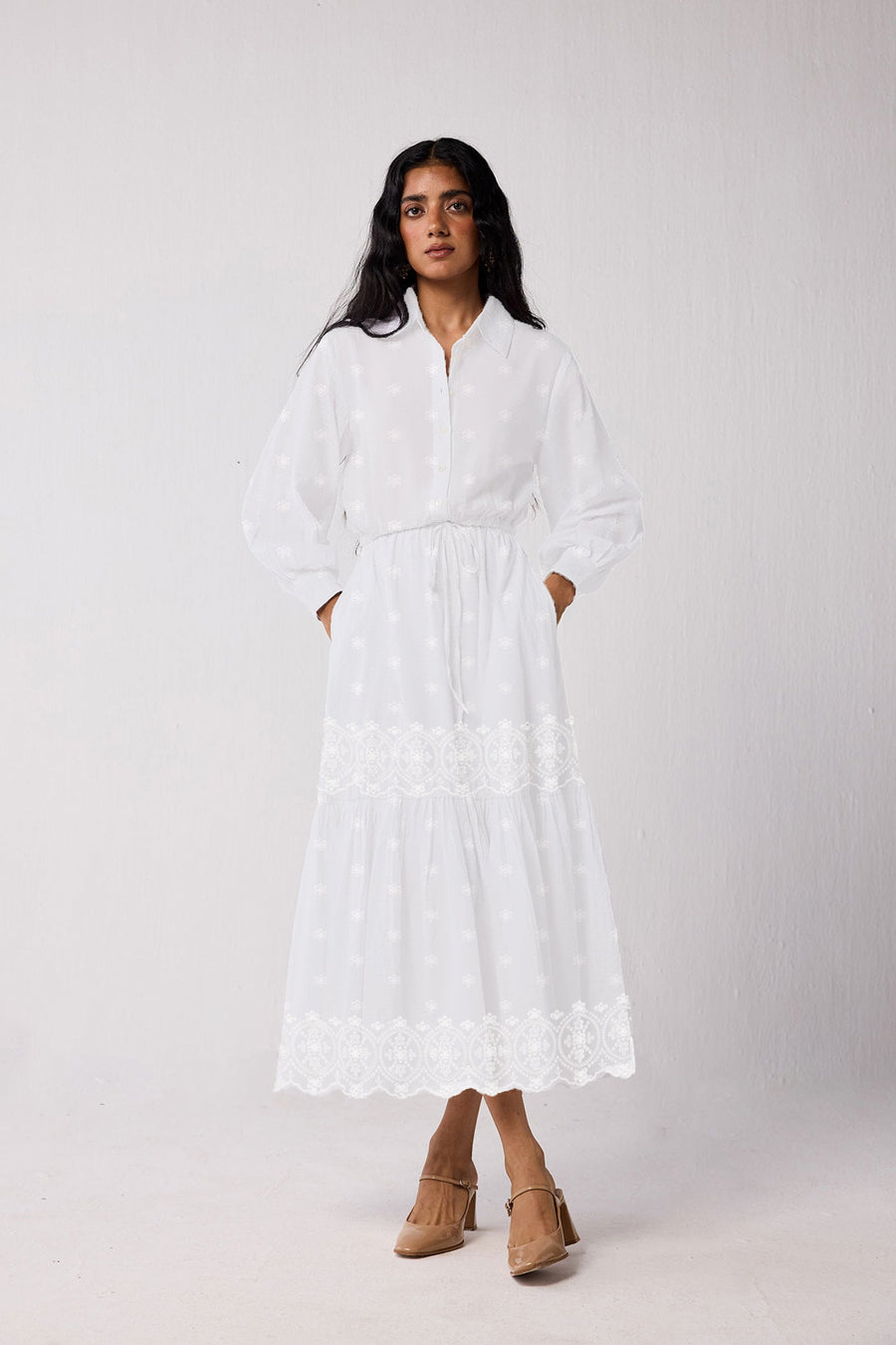 Full-body studio shot of a model presenting the white embroidered maxi tiered dress, emphasizing its relaxed vintage silhouette and functional side pockets, styled with tan pointed-toe heels.