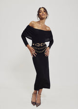 Full-length view of a model styling the off-the-shoulder geometric black knit cowl neck dress with an adjustable western double buckle belt, layered gold chain belt, and leopard print heels.