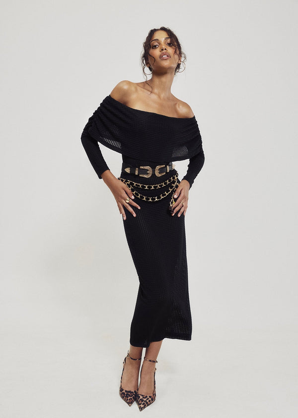 Full-length view of a model styling the off-the-shoulder geometric black knit cowl neck dress with an adjustable western double buckle belt, layered gold chain belt, and leopard print heels.