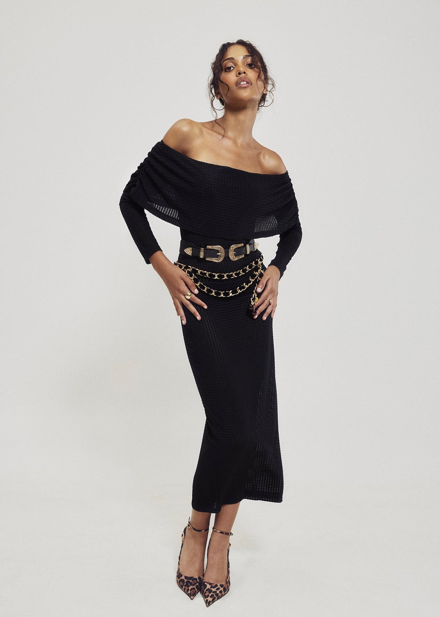 Full-length view of a model styling the off-the-shoulder geometric black knit cowl neck dress with an adjustable western double buckle belt, layered gold chain belt, and leopard print heels.