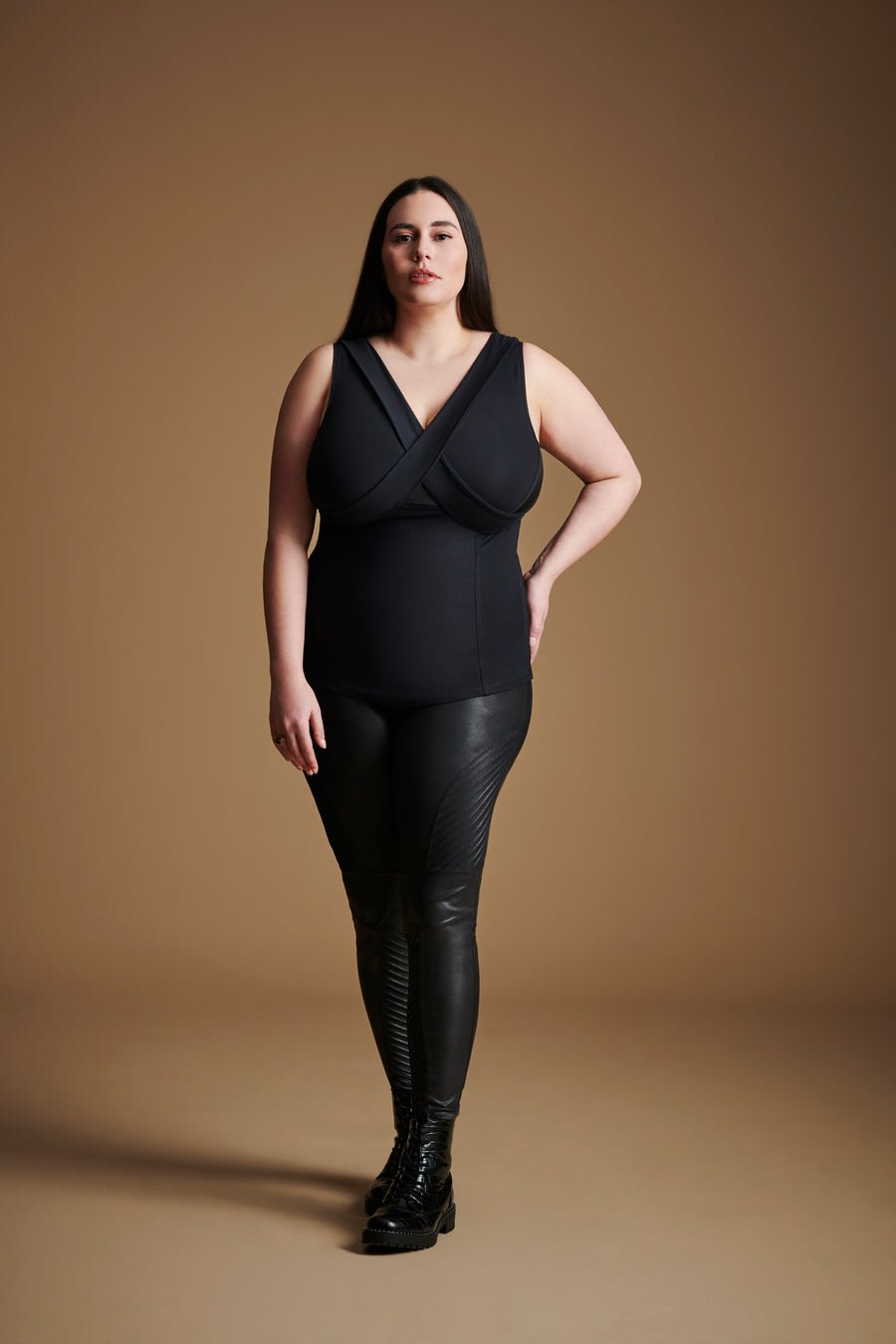 A woman in black Embrago leggings and top stands  against a brown background, showcasing a modern, stylish look.