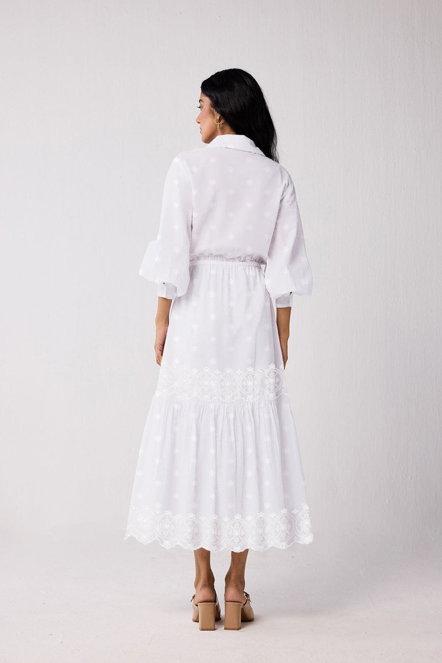Back view of a model walking in the white embroidered cotton dress, highlighting the clean shoulder yoke line and flowy tiered midi-to-maxi length drape.