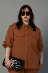 Dark-haired model with sunglasses wearing the terracotta Payton shirt jacket paired with a black woven leather clutch bag.