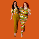 Full-body action shot of two models laughing against an orange background in the Blue Platypus Earth-toned Meadow Print jumpsuit and shirt.