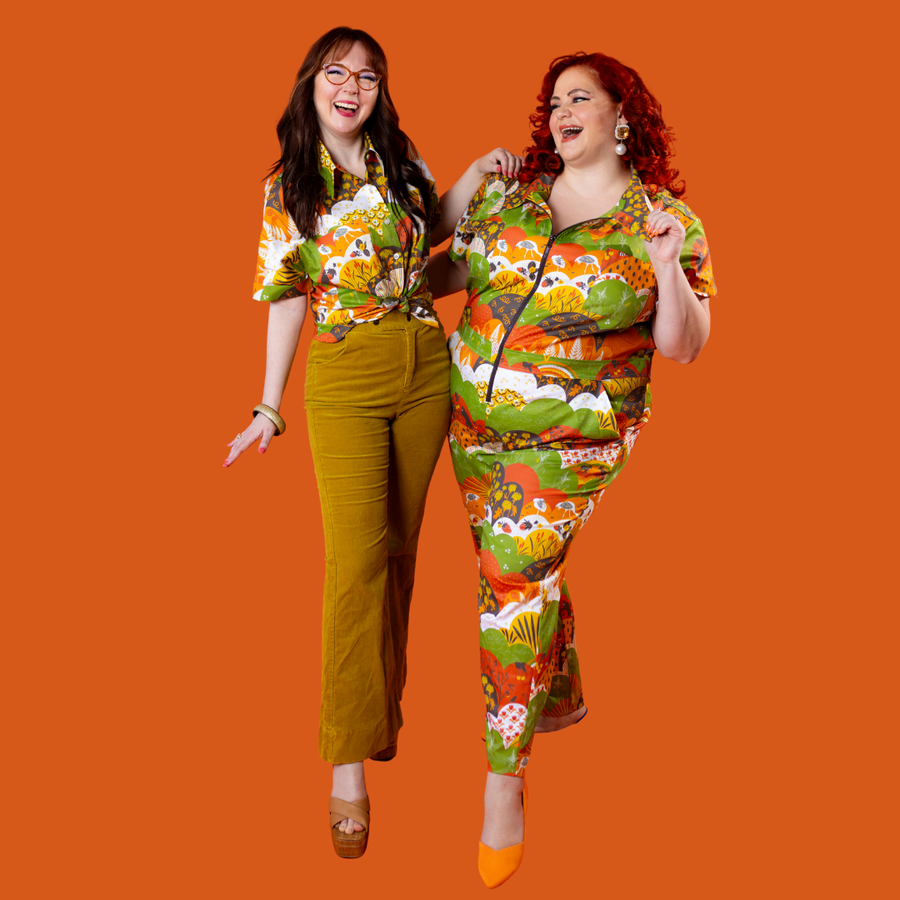 Full-body action shot of two models laughing against an orange background in the Blue Platypus Earth-toned Meadow Print jumpsuit and shirt.