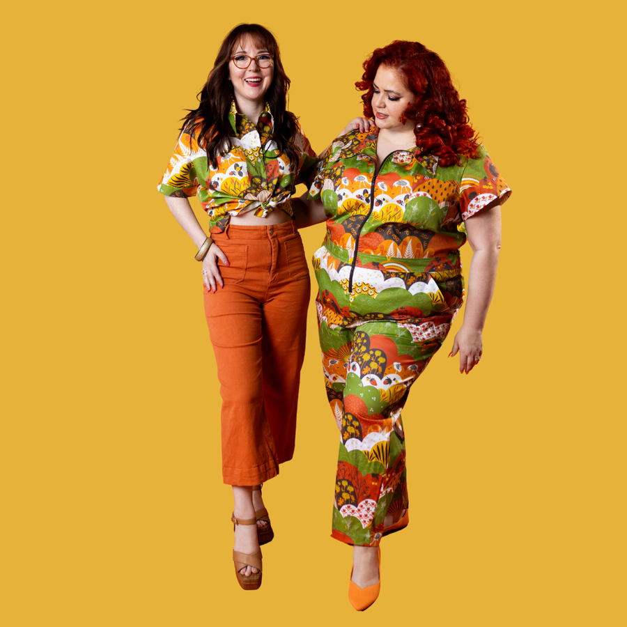 Full-body shot against a yellow background featuring two models wearing the Earth-toned Meadow Print jumpsuit and matching button-up shirt.