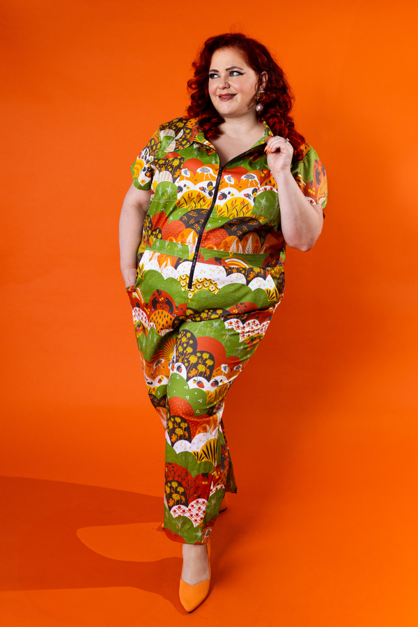Full-length shot of a red-haired model posing dynamic on an orange background wearing the Blue Platypus Jumpsuit in Earth-toned Meadow Print.