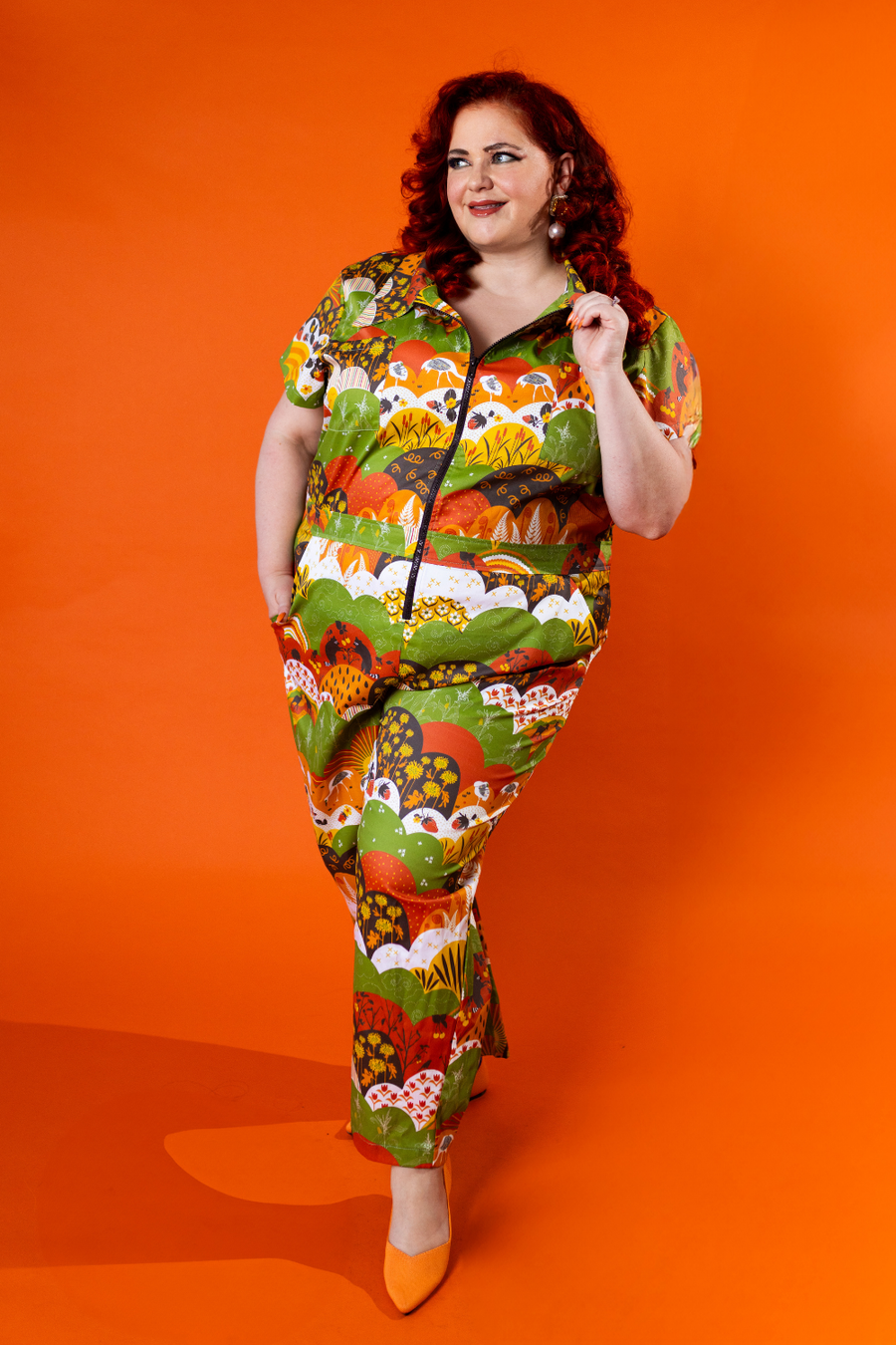Full-length shot of a red-haired model posing dynamic on an orange background wearing the Blue Platypus Jumpsuit in Earth-toned Meadow Print.