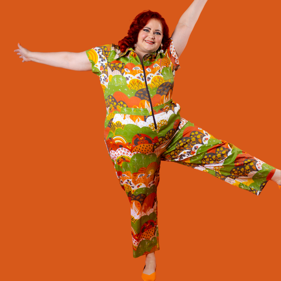 Full-length shot of a red-haired model posing dynamic on an orange background wearing the Blue Platypus Jumpsuit in Earth-toned Meadow Print.