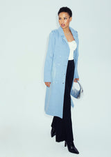 A model showcases the clean-front silhouette of the light sky blue Tamika denim trench coat styled open with black trousers and a metallic silver handbag.