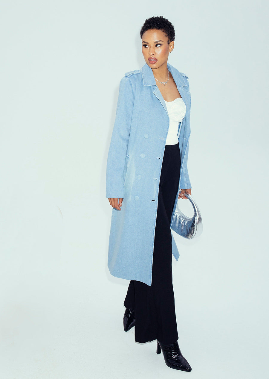 A model showcases the clean-front silhouette of the light sky blue Tamika denim trench coat styled open with black trousers and a metallic silver handbag.