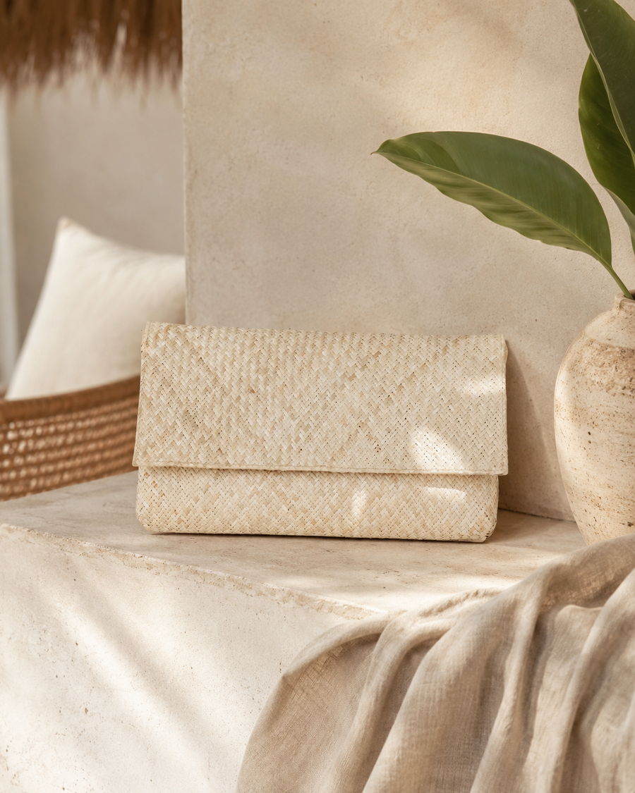 The natural woven clutch displayed horizontally on a minimalist stone console next to a clay vase and tropical green leaf.