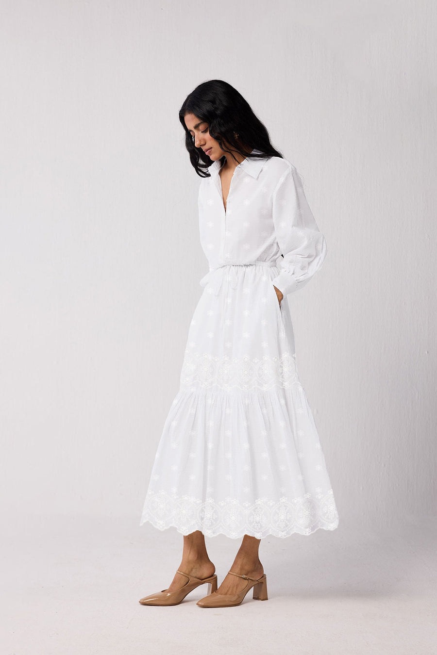 Side profile view of a model standing with her hand in her pocket, showcasing the effortless drape of the white organic cotton fabric and the neat wrist cuff details on the bishop sleeves.