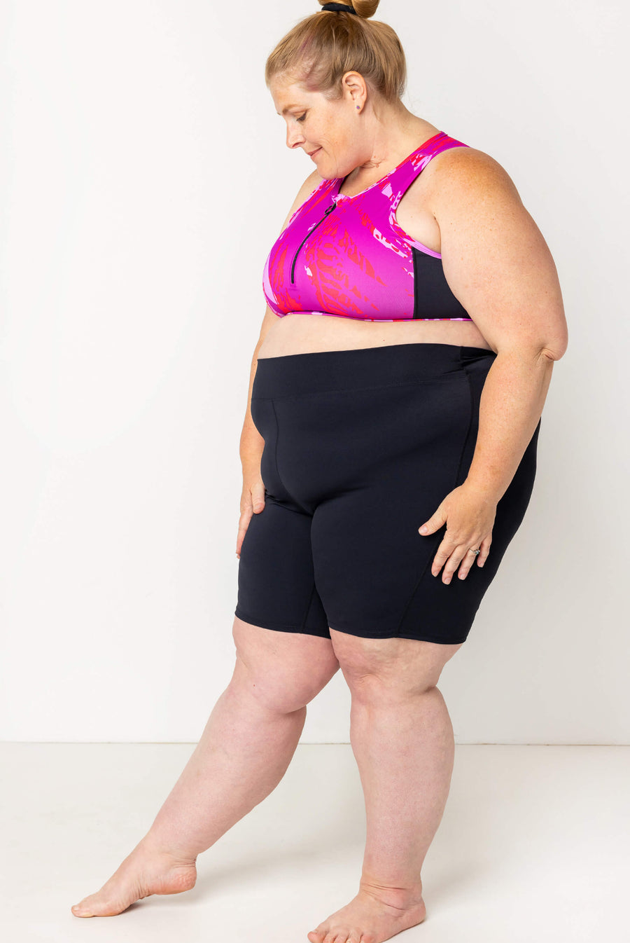 Full-body side profile of a blonde model wearing high-waisted black 9-inch bike-style swim shorts paired with a pink zip-front swim top.