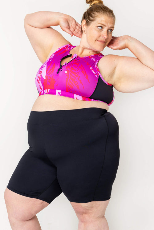 Three-quarter shot of a blonde model posing with hands behind her head wearing high-waisted black 9-inch swim shorts and a pink printed swim top.