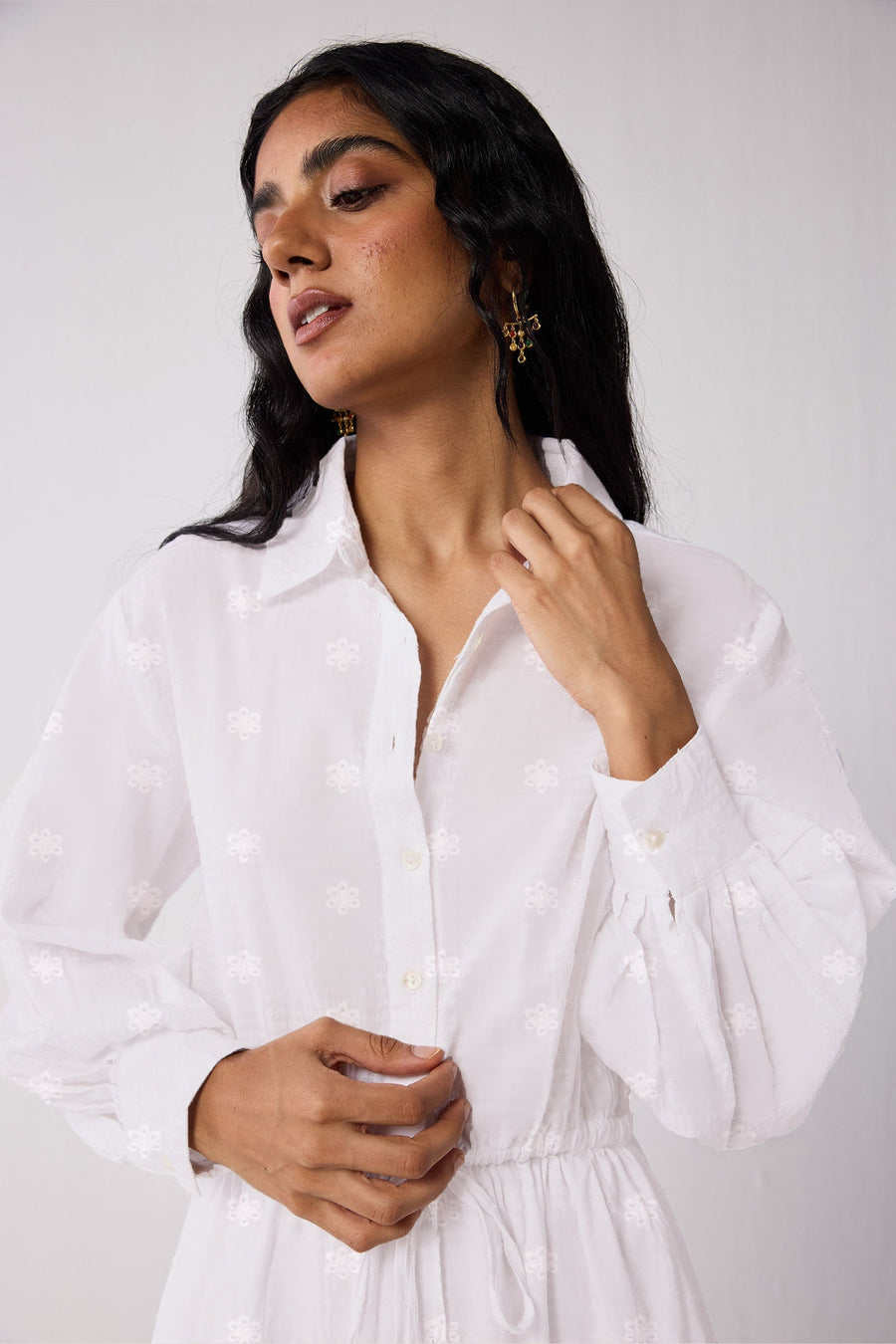 Close-up shot of a model adjusting the collar of the white Reistor dress, showcasing the intricate all-over floral embroidery motifs, the front button placket, and the structured fabric texture.