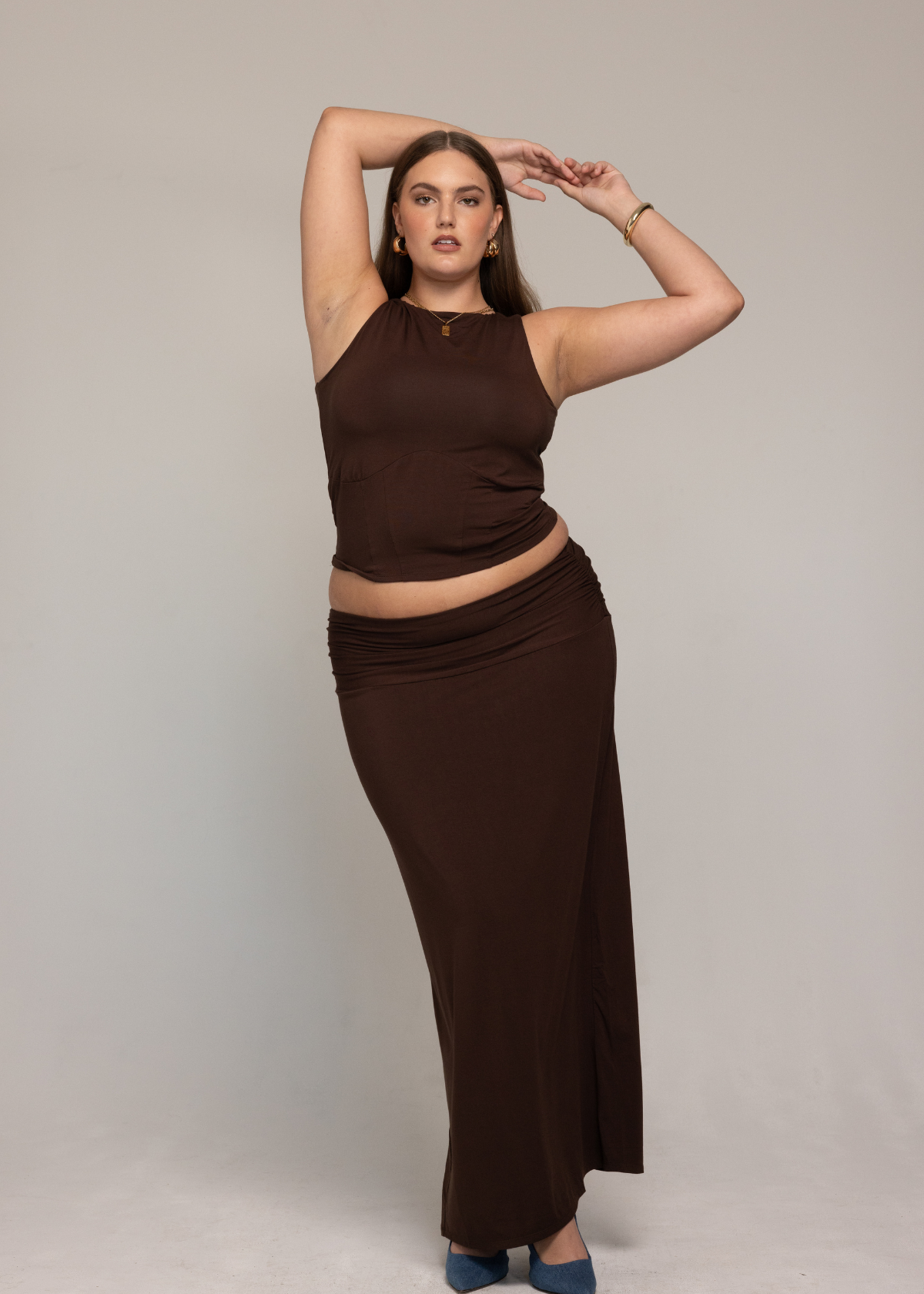 irl. They have one arm raised above their head and are sporting blue heels. Their makeup is polished, complemented by hoop earrings and bracelets.
