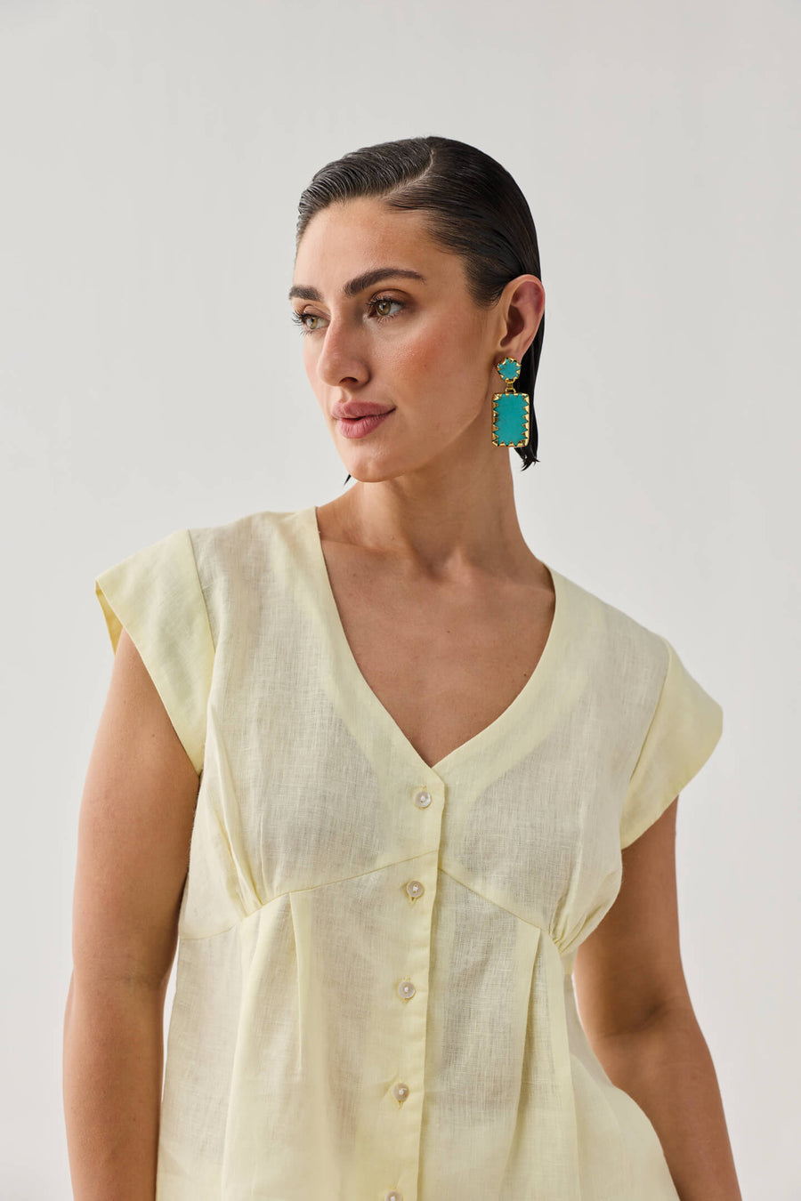 Studio portrait of a model looking over her shoulder while wearing the pale yellow Reistor linen top, accessorized with vibrant turquoise statement earrings.