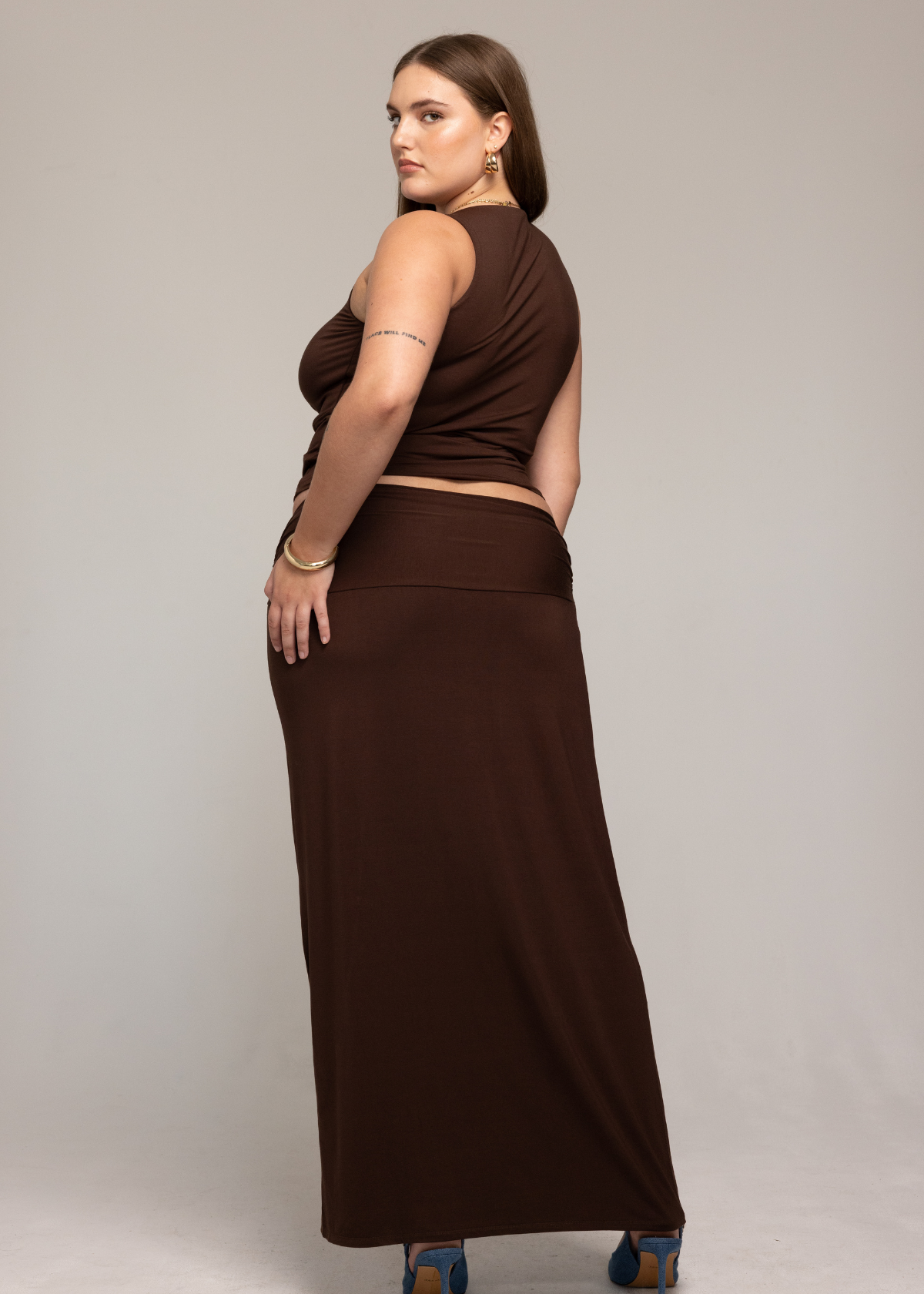 irl Valentina Skirt with a matching brown sleeveless top, paired with blue high heels. Their hair is worn down, and they have visible tattoos on their arms. The background is plain and light-colored.