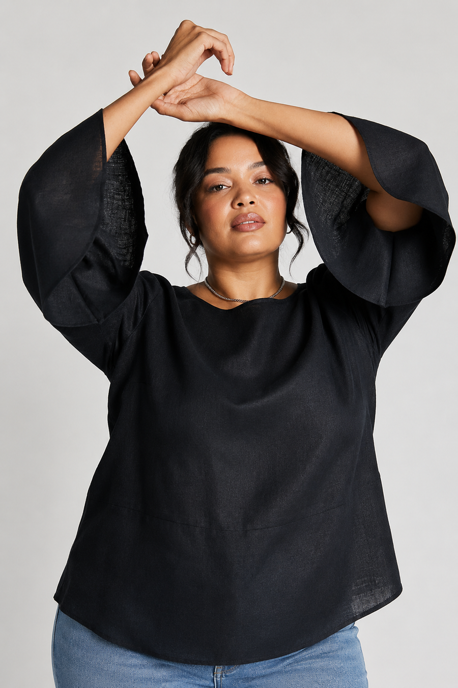Close-up shot of a plus-size model raising her arms above her head, beautifully showcasing the dramatic volume and fluid drape of the fit-and-flared bell sleeves on the black hemp top.