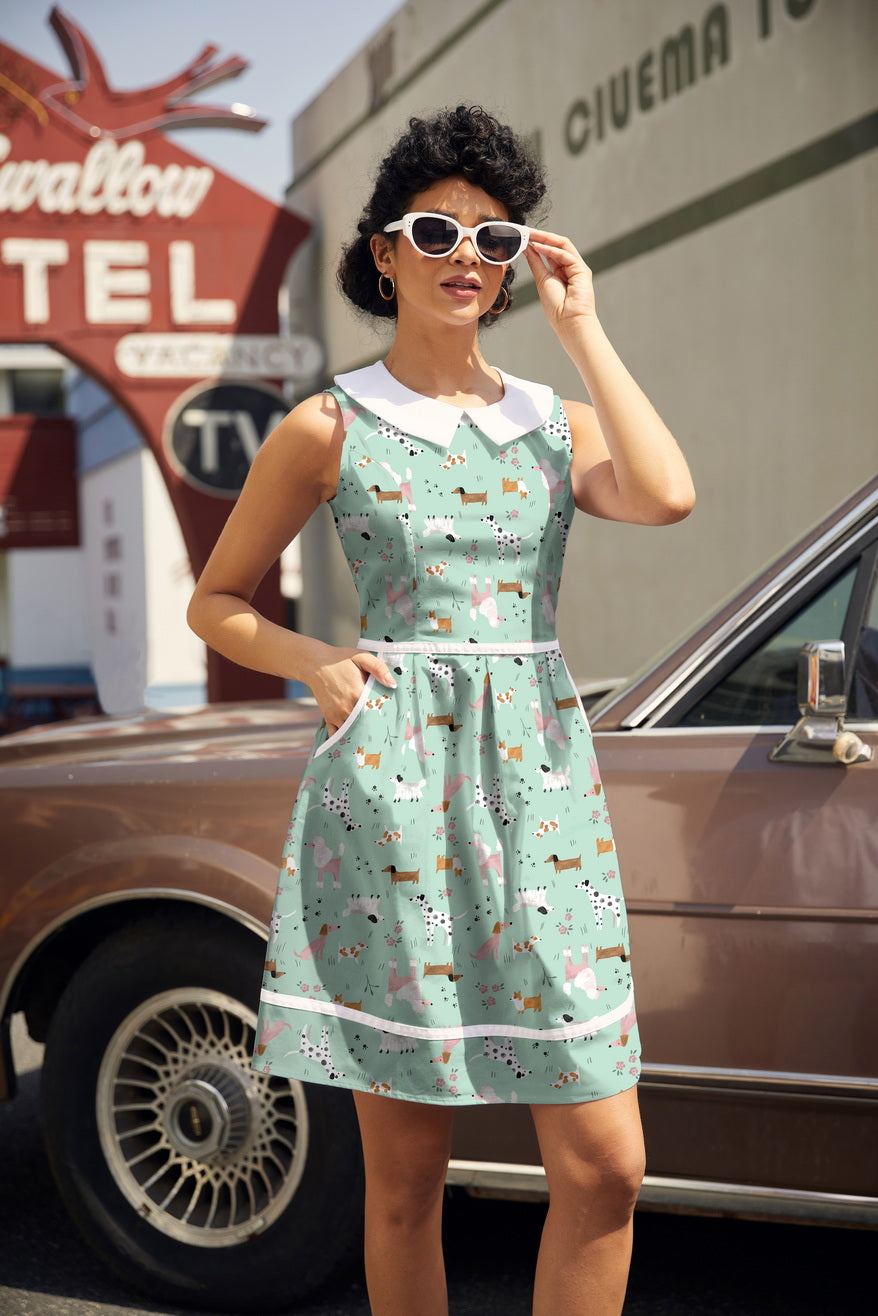 Model posing outdoors near a vintage car wearing the Retrolicious Dogs Collared Dress featuring a mint green dog print, white Peter Pan collar, and contrast piping.