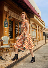Full-length photo of a model walking outside a cafe wearing the orange checkered cotton candy print Greta dress styled with black lace-up ankle boots.