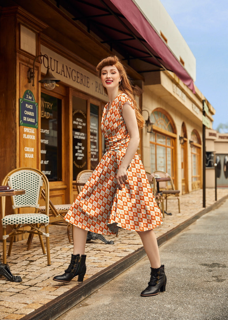 Full-length photo of a model walking outside a cafe wearing the orange checkered cotton candy print Greta dress styled with black lace-up ankle boots.
