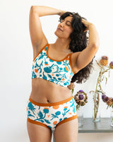 Front view studio shot of a model with long dark wavy hair running a hand through her hair, wearing the Robben's Artichokes teal and rust organic cotton longline bra set.