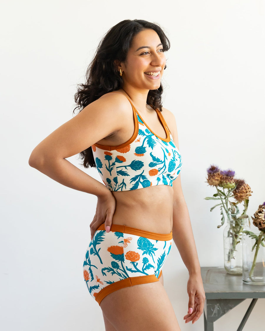 Side profile view of a model smiling while wearing the Robben's Artichokes longline bra with rust trim, standing next to dried artichoke flowers.