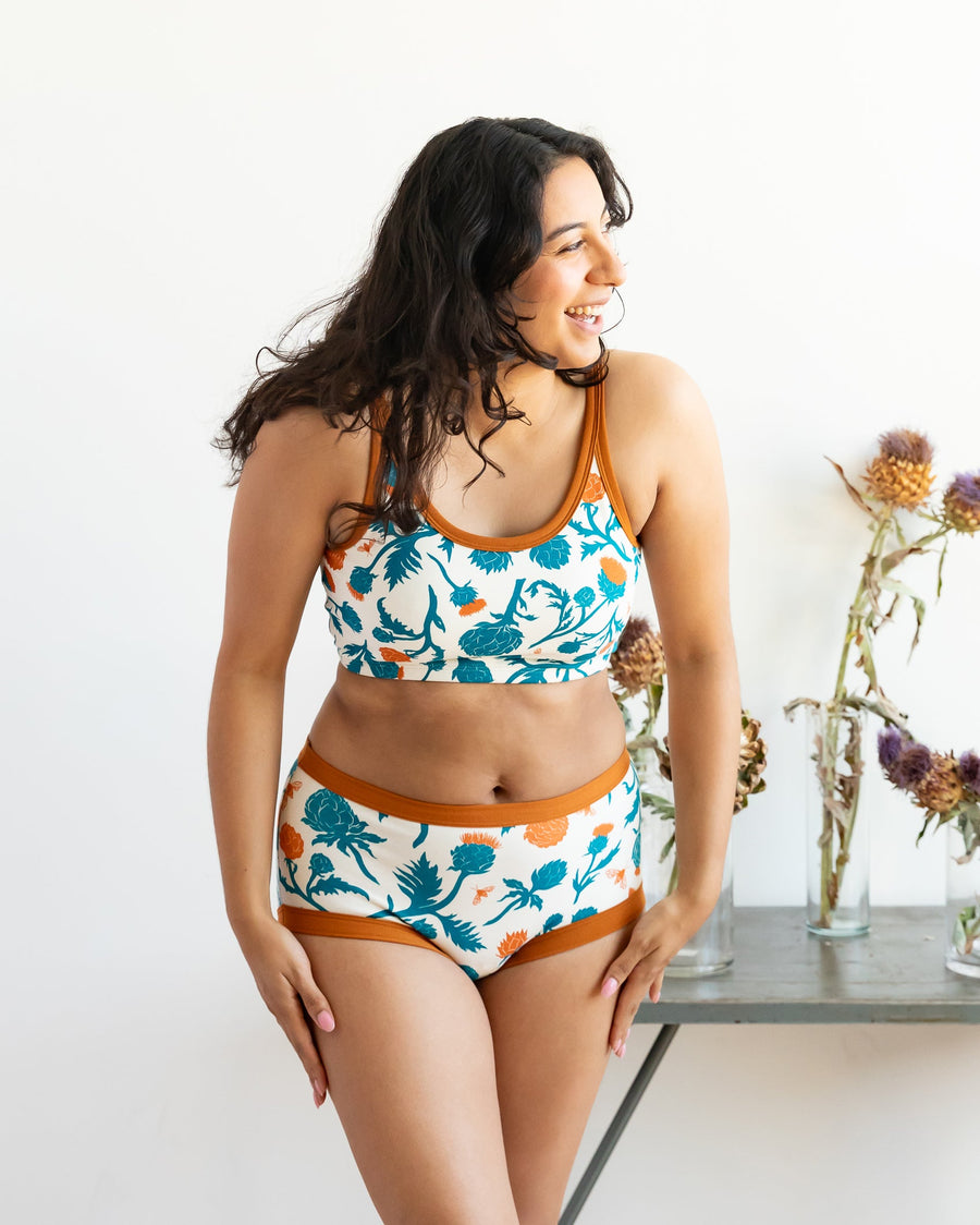 Front view studio shot of a model with long dark wavy hair running a hand through her hair, wearing the Robben's Artichokes teal and rust organic cotton longline bra set.
