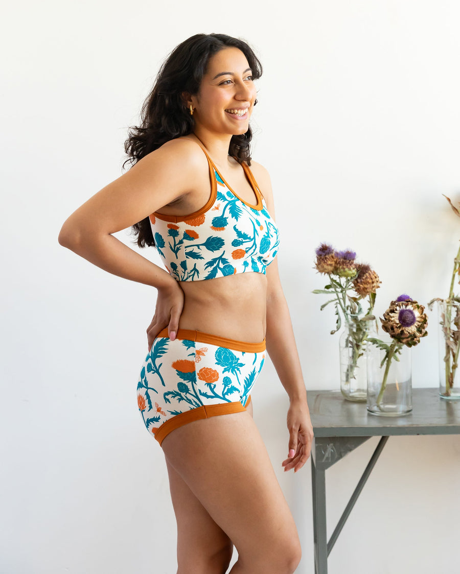 Side profile view of a model smiling while wearing the Robben's Artichokes longline bra with rust trim, standing next to dried artichoke flowers.
