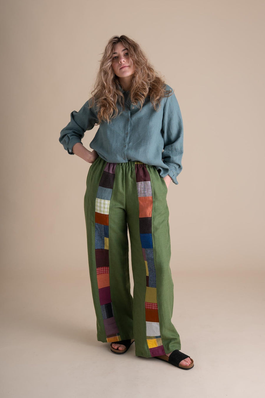 Full-length studio portrait of a curly-haired model wearing the handmade Joystripe Pants in Clover green paired with a teal button-down shirt.