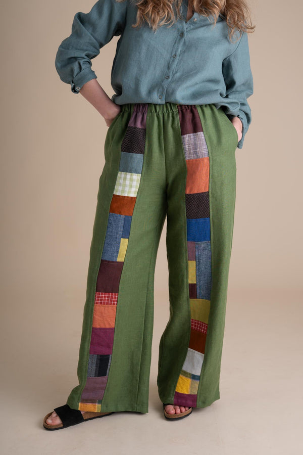 Front cropped view of a model wearing wide-leg green flax linen Joystripe Pants featuring colorful patchwork down each leg, styled with black sandals.