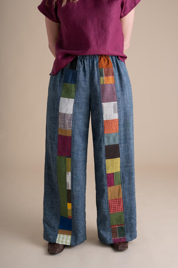 Front cropped view of a model wearing wide-leg indigo chambray Joystripe Pants featuring colorful patchwork down each leg and a soft elastic waistband.