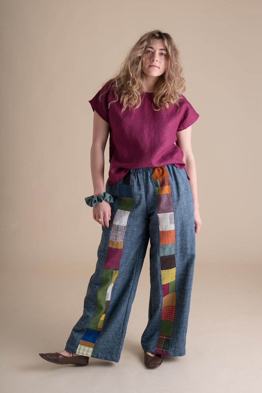 Full-length studio portrait of a model wearing handmade Joystripe Pants in Indigo Chambray with colorful patchwork details, styled with flat brown shoes.