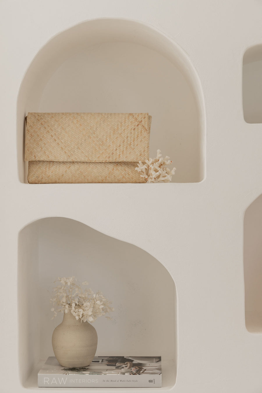 The natural variant of the handwoven clutch beautifully styled inside a clean white arched wall niche alongside a decorative coral branch.