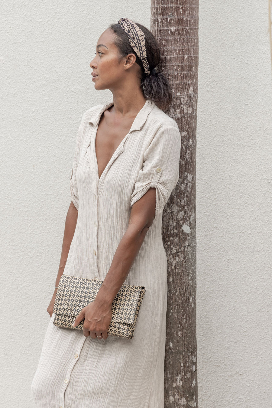 A model leans against a palm tree holding the geometric black motif woven straw clutch bag, paired with a relaxed neutral summer dress.