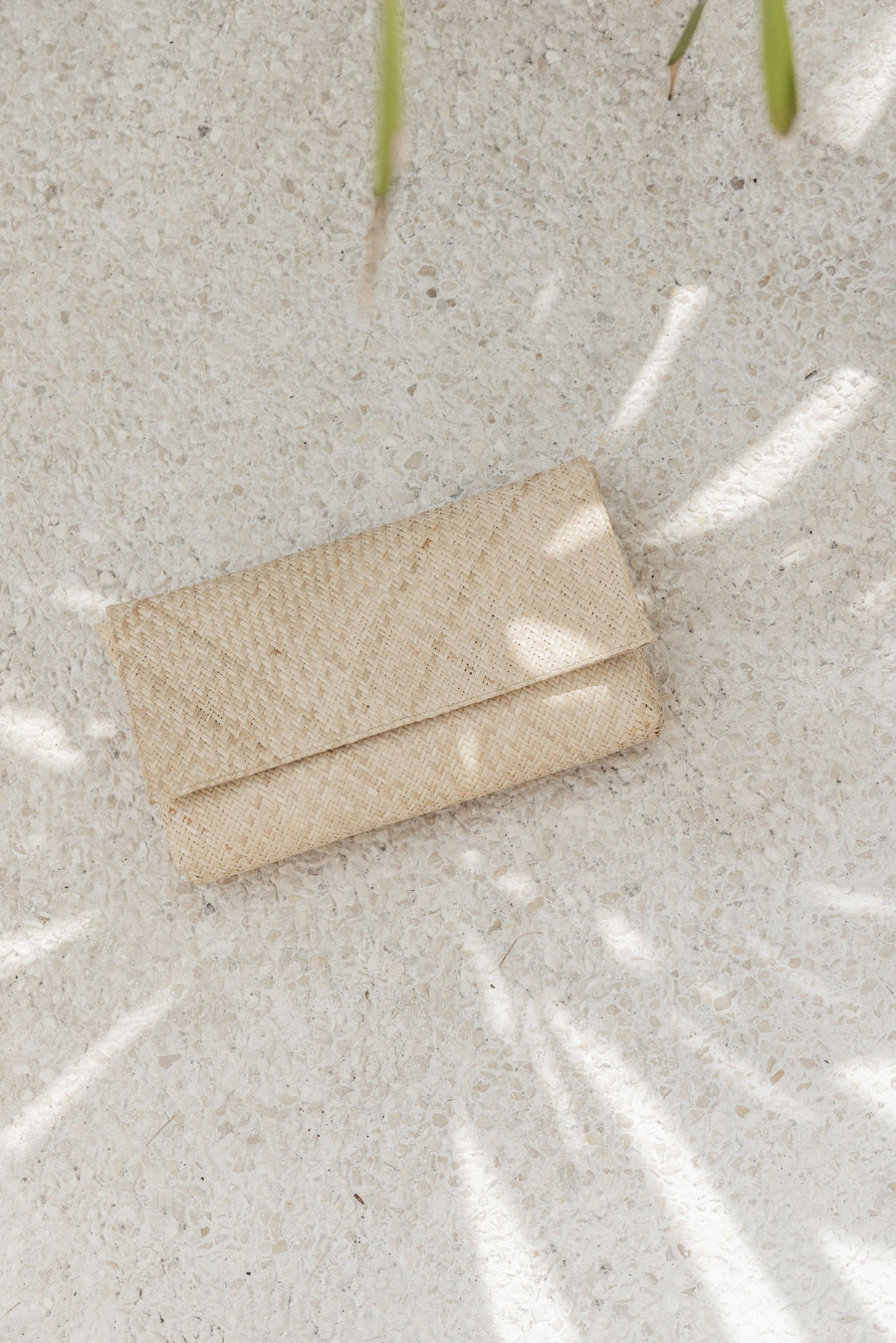 Top-down overhead view of the natural woven clutch resting on a speckled beige concrete background with soft tropical tree shadows.