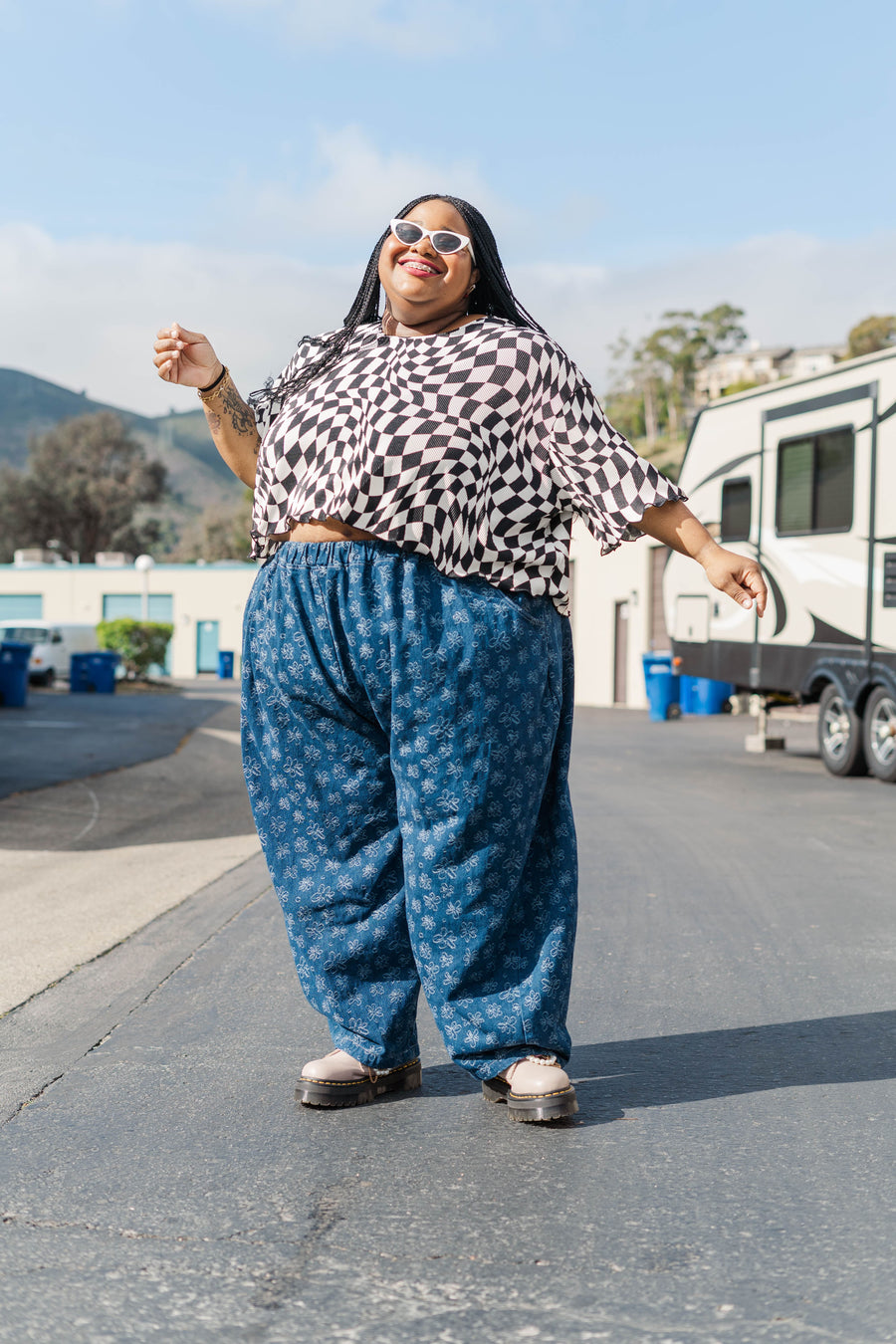 Outdoor full-length portrait of model Keda posing in the cropped Dizzy Favorite Tee in black and white wiggly checker print with cat-eye sunglasses.