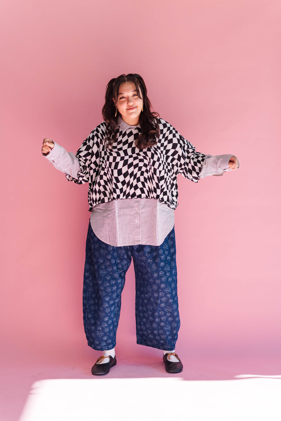 Full-length studio photo of model Angie wearing the Dizzy Favorite Tee layered over a striped button-down shirt paired with blue printed pants against a pink backdrop.